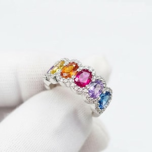 May include: A silver ring featuring a row of oval-cut gemstones in a rainbow of colors: yellow, orange, pink, purple, and blue. Each gem is surrounded by small, clear accent stones.