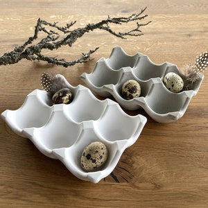 Könnte beinhalten: Zwei hellgraue, eiförmige Keramikschalen, die jeweils gefleckte Wachteleier und dekorative Federn enthalten. Die Schalen stehen auf einer Holzoberfläche, mit einem trockenen Ast im Hintergrund. Die Schalen haben sechs Eierfächer.