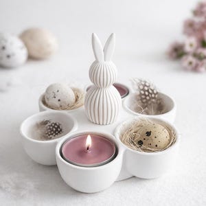 Könnte beinhalten: Ein weißes Keramik-Oster-Arrangement mit einer Hasenfigur, sechs kleinen Schalen und einer brennenden Kerze. Die Schalen enthalten gesprenkelte Eier, Federn und Stroh. Ein dekorativer Artikel für Ostern.
