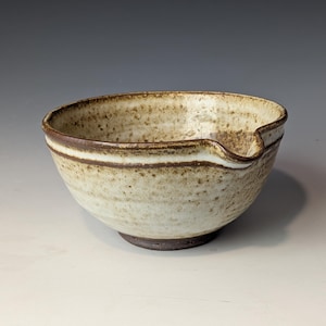 May include: A handmade ceramic bowl with a light beige speckled glaze and a dark brown rim. The bowl has a unique spout and a dark brown base. The bowl is approximately 6 inches in diameter.