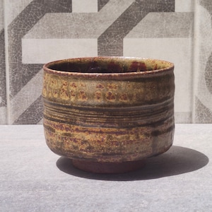 May include: A brown and green ceramic bowl with a textured surface and a wide rim. The bowl has a slightly flared base and is decorated with horizontal lines.