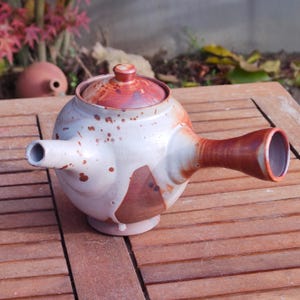 Può includere: Teiera in ceramica fatta a mano con corpo rotondo, coperchio e lungo beccuccio. La teiera presenta una glassa bianca e marrone macchiata con accenti marrone rossiccio. Il design è rustico e artistico.