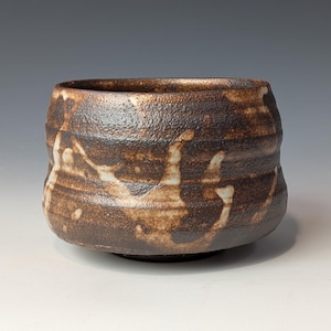 May include: A handmade, textured ceramic bowl with a dark brown exterior and lighter brown and white accents. The bowl has a rounded shape with a slightly irregular rim and a small, flat base. The pottery has a rustic, organic aesthetic.