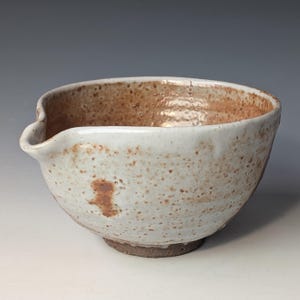 May include: A handmade ceramic bowl with a spout, featuring a speckled white and brown glaze. The bowl has a rounded shape with a slightly flared rim and a dark brown base. The interior has a darker brown color.