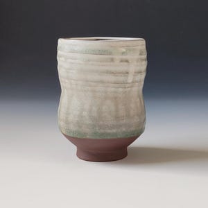 Może przedstawiać: Biały ceramiczny kubek z zieloną glazurą i brązową podstawą. Kubek ma lekko rozszerzający się rant i zaokrąglone ciało. Glazura jest nierówna, tworząc teksturalną powierzchnię.