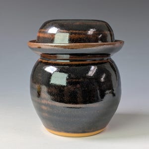 May include: A ceramic jar with a matching lid, featuring a glossy dark brown and black glaze with subtle horizontal bands of lighter brown. The vessel has a rounded body and a wide, flat-rimmed lid that sits securely on top of the container.