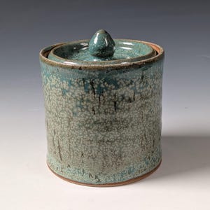 Może przedstawiać: Cylindryczny ceramiczny pojemnik z pokrywką wyposażoną w mały uchwyt w kształcie gałki. Zewnętrzna strona pokryta jest nakrapianym szkliwem w odcieniach turkusu, błękitu i bieli, o teksturze z widocznymi ciemnymi pionowymi smugami na powierzchni.