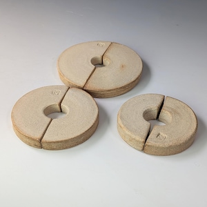 May include: Three circular, beige ceramic objects with a textured, matte finish. Each piece features a central hole and a single straight slit extending from the centre to the outer edge. A small, square maker's mark is stamped on each piece.