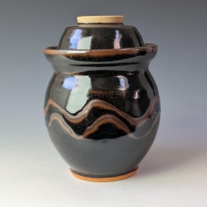 Op de afbeelding: Een keramische pot met deksel, voorzien van een glanzend zwart glazuur en een horizontaal golvend bruin bandpatroon. De pot heeft een afgerond lichaam, een uitlopende rand en een ongeglazuurde beige basis en handvat op het deksel.