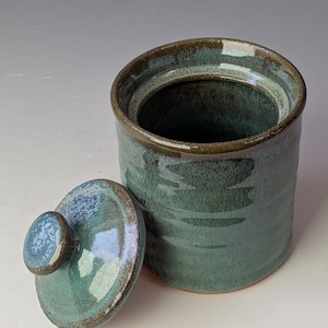 Könnte beinhalten: Ein zylindrisches Keramikgefäß mit einer glänzenden, gesprenkelten Glasur in Blaugrün und Dunkelgrün. Das Gefäß verfügt über einen passenden abnehmbaren Deckel mit einem kleinen Knaufgriff und zeigt subtile Farbvariationen sowie eine glatte Oberfläche.
