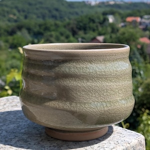 May include: An olive-green ceramic bowl with a crackled glaze. The bowl has a rounded shape with a flared rim and sits on a small, unglazed base. The exterior features subtle, horizontal ridges. A handmade piece.