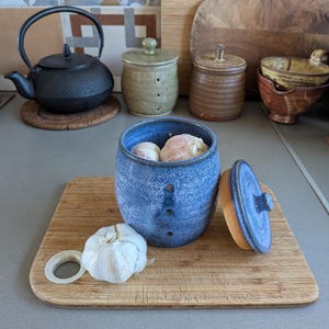 以下が含まれることがあります： 斑点のある釉薬と垂直の通気孔が特徴の、蓋付きの青い陶器製ガーリックキーパー。容器にはニンニクが2個入っており、もう1個のニンニクが容器の横に置かれています。