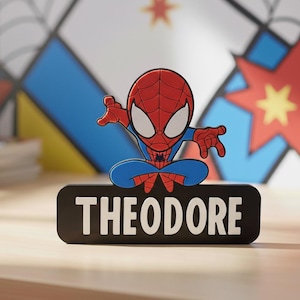 May include: A Spider-Man figure in a seated pose, with a red and blue costume, sits on a black nameplate with "THEODORE" in white. The background has a colourful comic book-style design.
