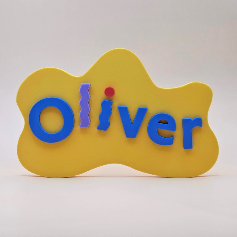 May include: A yellow cloud-shaped sign with the name "Oliver" spelled out in blue letters. The letters are raised and have a 3D effect. There is a small red circle and a purple zig-zag pattern on the sign.