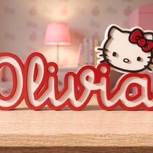 May include: A red and white name sign reading "Olivia" with a Hello Kitty design. The sign features a red outline and white interior. The Hello Kitty character has a red bow. The sign is made of layered materials.