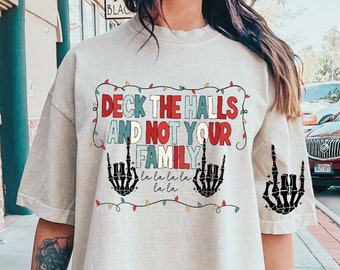 May include: A white t-shirt with a Christmas-themed graphic that says "Deck the halls and not your family" with skeleton hands giving the middle finger and a string of Christmas lights around the text.