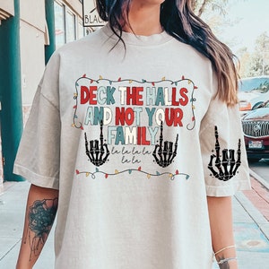 Puede incluir: Una camiseta blanca con un gráfico navideño que dice "Deck the halls and not your family" con manos de esqueleto haciendo el dedo medio y una cadena de luces navideñas alrededor del texto.