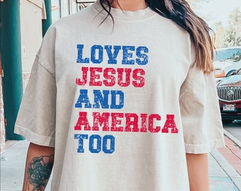 Op de afbeelding: Een licht beige T-shirt met een verweerde grafische print met de tekst "Loves Jesus and America Too" in rode en blauwe letters.