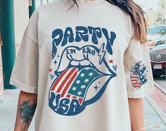 May include: A light beige oversized t-shirt with a graphic print of a red, white, and blue tongue sticking out of a mouth with the text "Party in the USA" in blue. The graphic has a vintage, distressed look.