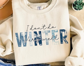 Puede incluir: Una sudadera de color crema con el texto "I don't do winter very well" en letras azules y blancas con un aspecto desgastado. La sudadera está colocada sobre una superficie de madera con un sombrero marrón, unos vaqueros azules y una pequeña planta en maceta.