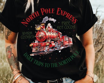 May include: Black t-shirt featuring a festive illustration of a red steam train with the text "North Pole Express." The design includes the phrases "Warm Cookies," "Free Hot Cocoa," and "Daily Trips to the North Pole."