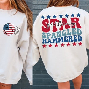 May include: White sweatshirt with a red, white, and blue smiley face with peace sign on the front and the text "getting STAR SPANGLED HAMMERED" with red, white, and blue stars on the back.
