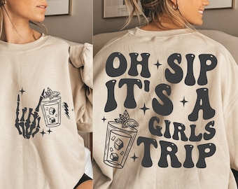 Könnte beinhalten: Ein beiger Pullover mit einem grafischen Druck einer Skeletthand, die ein Glas mit einem Getränk mit einer Zitronenscheibe und einem Minzblatt hält. Der Text "OH SIP IT'S A GIRLS TRIP" ist auf der Rückseite des Pullovers aufgedruckt.