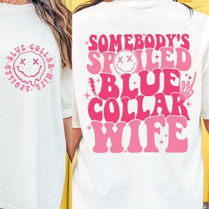 May include: White t-shirt with pink text that reads "Somebody's Spoiled Blue Collar Wife" with a smiley face graphic. The back of the shirt has a smaller smiley face graphic with the text "Spoiled Blue Collar Wife".