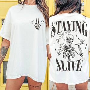 May include: Off-white t-shirt with a skeleton hand making a peace sign on the front. The back features a skeleton holding a coffee cup with the words "STAYING ALIVE" in black. The shirt has a relaxed fit.