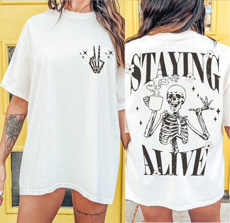 May include: Off-white t-shirt with a skeleton hand making a peace sign on the front. The back features a skeleton holding a coffee cup with the words "STAYING ALIVE" in black. The shirt has a relaxed fit.