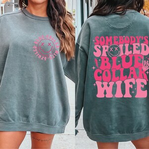 May include: A dark green sweatshirt with a pink graphic that says "Somebody's Spoiled Blue Collar Wife" with a smiley face and a skeleton hand.