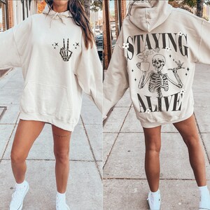 May include: Beige hoodie with a front pocket and a hood. The front features a black skeleton hand making a peace sign. The back of the hoodie has a skeleton graphic with the words "STAYING ALIVE".