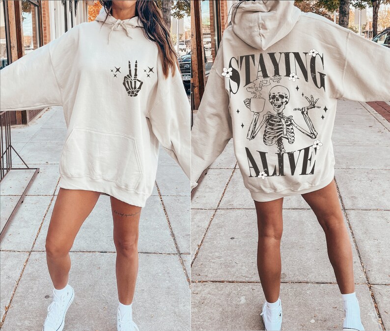 May include: Beige hoodie with a front pocket and a hood. The front features a black skeleton hand making a peace sign. The back of the hoodie has a skeleton graphic with the words "STAYING ALIVE".