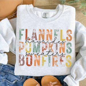 Peut inclure: Un sweat-shirt gris avec un design graphique de style vintage représentant les mots "Flannels, Hayrides, Pumpkins, Sweaters, Bonfires" en lettres colorées et vieillies.