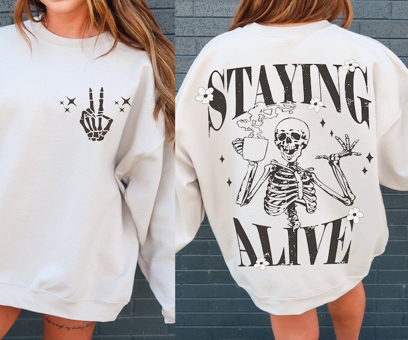 May include: Cream-colored sweatshirt with a black skeleton peace sign graphic on the front and the words "STAYING ALIVE" with a skeleton and coffee cup graphic on the back. The design includes stars and flowers.
