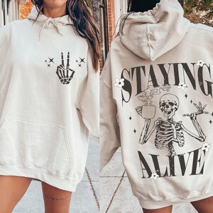 May include: Beige hoodie with a front pocket and drawstring hood. The front features a black skeleton hand making a peace sign. The back displays a skeleton holding a coffee cup with the words "STAYING ALIVE" in black.