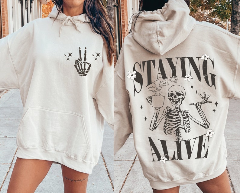 May include: Beige hoodie with a front pocket and drawstring hood. The front features a black skeleton hand making a peace sign. The back displays a skeleton holding a coffee cup with the words "STAYING ALIVE" in black.