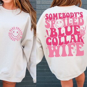 May include: White sweatshirt with pink text that reads "Somebody's Spoiled Blue Collar Wife" on the back. The front of the sweatshirt has a pink smiley face with a dripping mouth inside a circle with the text "Spoiled Blue Collar Wife" around it.