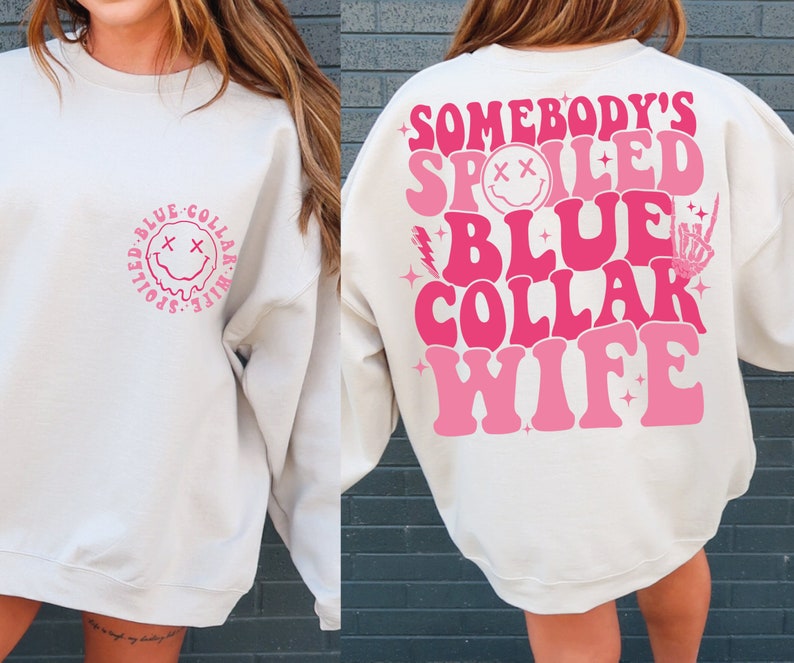May include: White sweatshirt with pink text that reads "Somebody's Spoiled Blue Collar Wife" on the back. The front of the sweatshirt has a pink smiley face with a dripping mouth inside a circle with the text "Spoiled Blue Collar Wife" around it.
