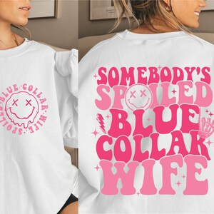 May include: White sweatshirt with pink text that reads "Somebody's Spoiled Blue Collar Wife" with a pink smiley face and other pink graphic elements.