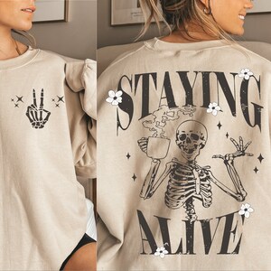May include: Beige sweatshirt with a skeleton peace sign on the front and the words "STAYING ALIVE" on the back. The back graphic features a skeleton holding a coffee cup, with floral and star accents.