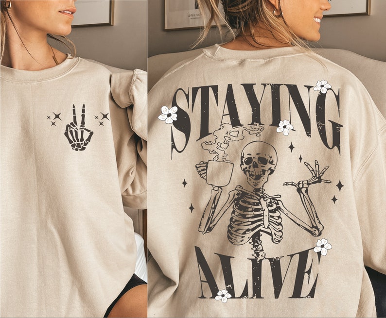 May include: Beige sweatshirt with a skeleton peace sign on the front and the words "STAYING ALIVE" on the back. The back graphic features a skeleton holding a coffee cup, with floral and star accents.