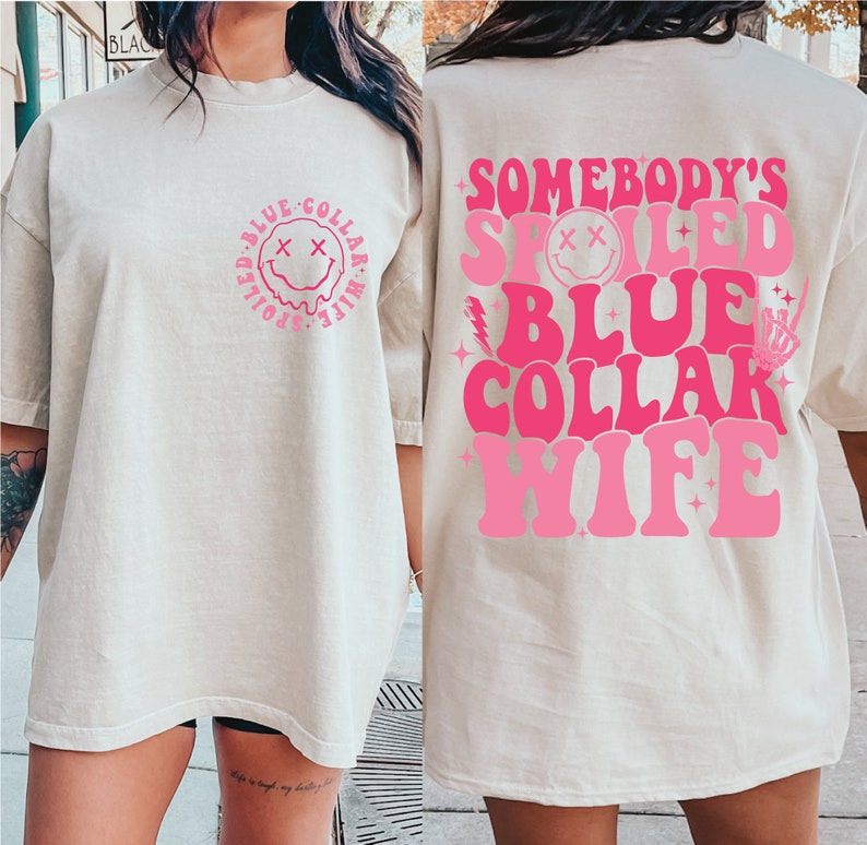 May include: An oversized t-shirt in a light beige colour with a pink graphic on the front and back. The front graphic is a smiley face with the text "Spoiled Blue Collar Wife" around it. The back graphic says "Somebody's Spoiled Blue Collar Wife" in a retro font.