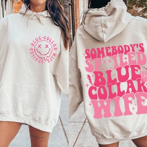 May include: A cream-coloured hoodie with a pink graphic that says "Somebody's Spoiled Blue Collar Wife" with a smiley face and other design elements.