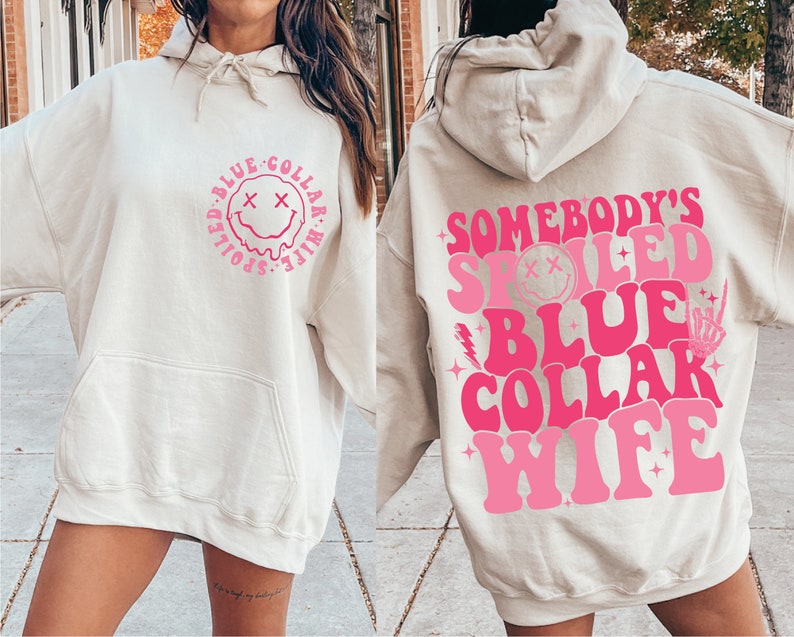 May include: A cream-coloured hoodie with a pink graphic that says "Somebody's Spoiled Blue Collar Wife" with a smiley face and other design elements.