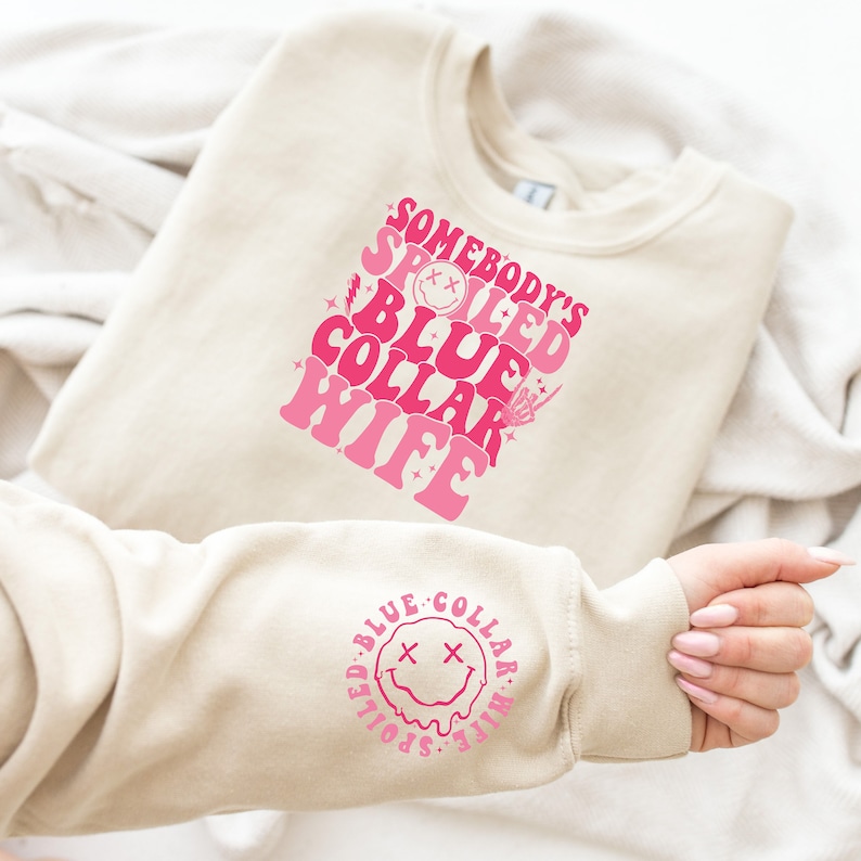 May include: A beige sweatshirt with a pink graphic that says "Somebody's Spoiled Blue Collar Wife" with a smiley face. The graphic is repeated on the sleeve of the sweatshirt.
