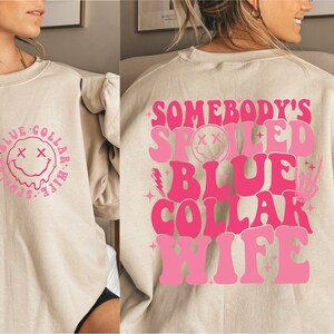 May include: A light beige sweatshirt with a pink graphic on the back that says "Somebody's Spoiled Blue Collar Wife" and a pink graphic on the front that says "Somebody's Spoiled Blue Collar Wife" with a smiley face.