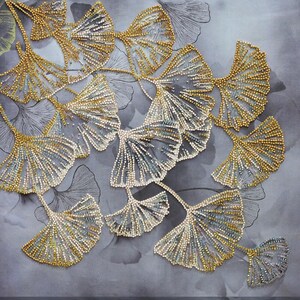 May include: Close-up of a textile featuring beaded ginkgo leaf designs in gold, silver, and white. The leaves are arranged on a gray background with subtle leaf patterns, creating a textured and elegant aesthetic. The intricate beadwork adds a touch of luxury.