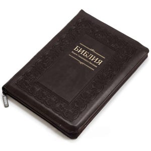 Pode incluir: Uma capa de livro com zíper em marrom escuro, apresentando uma borda decorativa em relevo e o texto dourado "БИБЛИЯ" seguido de "Книги Священного Писания" centralizado na capa frontal. O estojo retangular possui um puxador de zíper de metal.