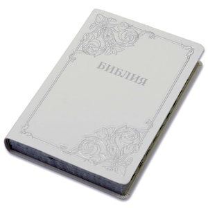 May include: A closed, cream-coloured book with the Cyrillic word "БИБЛИЯ" printed on the cover. The cover features ornate, silver-coloured floral corner designs and a silver border. The book's edges are grey.